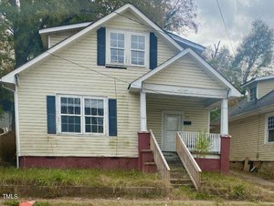 The small, two-story house at 436 N Chestnut Street in Henderson boasts pale yellow siding and dark shutters. It features a covered front porch with a white railing and red steps. The yard is overgrown with grass, and a neighboring home is partly visible to the right.