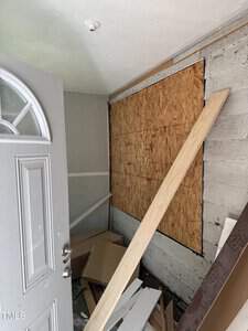 A cluttered room with an open door, showing building materials like plywood and wooden planks scattered on the floor. The walls are partially covered with wood panels. A beam leans against the wall.