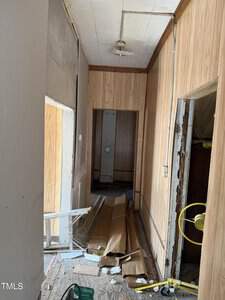 A narrow, unfinished hallway with wooden paneling and a partially exposed ceiling. The floor is cluttered with wooden planks and construction materials. A doorway at the end and an open door on the right lead to other unfinished rooms.