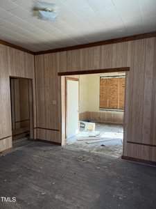 An empty room with wood-paneled walls and a dark, worn floor is shown. A doorway leads to another room with yellowed walls, scattered tools, and a boarded window, indicating renovation work. A ceiling light fixture is visible.