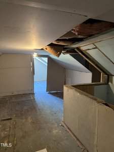 An unfinished attic with damaged drywall and exposed insulation on the ceiling. The room is lit by a single bulb, and there's a small window at the far end. The floor is wooden and appears worn. Several slanted walls are in view.