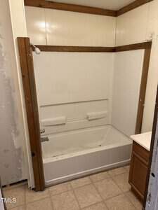 A small bathroom with a white bathtub and shower combination installed in a wooden-framed alcove. The walls are plain, and there is a partial view of a small vanity with a countertop on the right. The floor is tiled.
