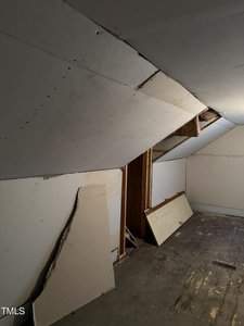 An attic space with partially installed drywall on the slanted ceiling. Some pieces of drywall are stacked against the wall, and a dismantled door is leaning on the right side. The floor appears unfinished or worn.