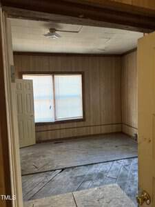 A small, unfurnished room with wood-paneled walls and a window with closed blinds. The floor is partially covered with plywood, and a ceiling fan is installed. The room appears in need of renovation.