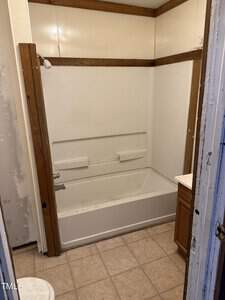 A small bathroom with a bathtub and showerhead, surrounded by beige walls and a wooden frame. The floor is tiled, and a wooden cabinet is partially visible on the right side. The room has a basic, unfinished appearance.