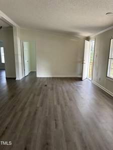 A bright, empty room with light gray laminate flooring and white walls. Sunlight enters through a window and an open door on the right side, leading outside. A doorway on the left wall opens into another room.