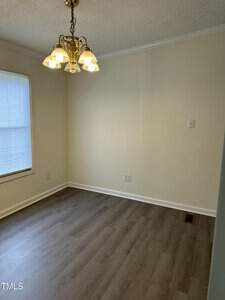 A small, empty room with light-colored walls and wooden flooring. It features a large window with blinds and a ceiling-mounted gold chandelier with five lights.