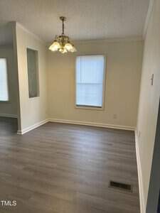 Empty room with light-colored walls and wood flooring. A window with blinds lets in natural light. A brass chandelier hangs from the ceiling, and a small decorative glass panel is set in a dividing wall.