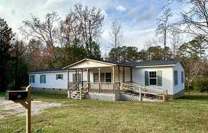 A white mobile home with a wooden ramp and porch sits on a grassy plot at 60 Casei Lane, Henderson, surrounded by trees. A mailbox bearing the number is visible on the left under the partly cloudy sky.