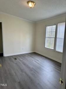 An empty room with light-colored walls, a single window with blinds, and wood-patterned flooring. A small ceiling light fixture is centered on the textured ceiling. A partially open door reveals a closet.