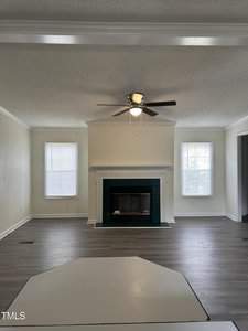 A living room with light-colored walls and wood flooring features a central fireplace framed by two tall windows. A ceiling fan with a light is mounted on the ceiling. The room appears spacious and empty.
