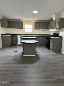 A modern kitchen with gray cabinets and a central island. The room features stainless steel appliances, a large window above the sink, and wood-look flooring. The backsplash is dark, contrasting with the light walls.