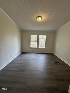 An empty room with light-colored walls and dark wooden flooring. A ceiling light fixture is centered, and two windows allow natural light to enter.