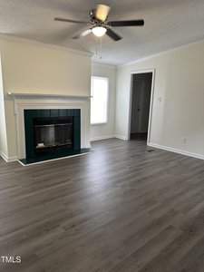 A living room with light gray hardwood floors, off-white walls, and a ceiling fan with lights. A fireplace with a dark green tile surround is centered on the left wall. A door and a large window are visible on the right side.