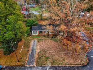 Aerial view of a brick house surrounded by autumn trees. The yard is covered in fallen leaves, and a driveway leads to the house. Nearby, other homes and streets can be seen amid the colorful foliage.
