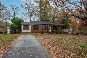 A small, single-story brick house with a porch, set in an autumn landscape. The yard is covered in fallen leaves, and the driveway leads up to the front door. Tall trees, mostly bare, surround the house.
