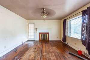 A vacant living room with hardwood floors and a ceiling fan. There are cream-colored walls, a white fireplace with a gold-rimmed firewood cover, and a red bucket in the corner. Purple curtains partially cover the large window on the right.