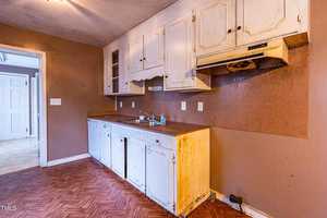 An old kitchen with white cabinets and brown walls. The countertop is worn, and there is a sink with a soap bottle nearby. The floor has a herringbone pattern. The room appears aged and in need of renovation.