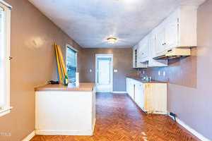 A narrow kitchen with brown walls, white cabinets, and a wooden floor. There's a small counter on the left with a few items. The room is lit by a ceiling light and a window. A door leads to another room at the end. The cabinets show signs of wear.