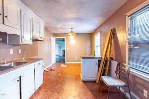 A dimly lit, cluttered kitchen with white cabinets and a single sink. The hardwood floor shows debris, and a wooden chair with boards leans against the wall by the window. A hanging light fixture is above, with a doorway leading to another room.