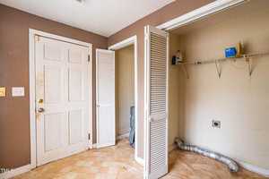 Empty laundry room with brown walls, a closed white door, folding closet doors, a wire shelf with cleaning supplies, and a dryer vent hose on the floor. The floor has a light checkered pattern.