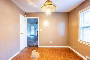 Empty room with tan walls and a wooden floor with a damaged area. A bright ceiling light hangs above. A doorway leads to another room with hardwood flooring and a window. Natural light enters through a window on the right.