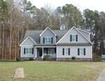 Located at 129 Creekside Drive in Oxford, this charming two-story gray house features white trim, a covered porch, and a gabled roof. It's nestled amid a grassy lawn, with a driveway leading to the entrance and trees providing a picturesque backdrop.