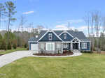 Located at 3831 Pickett Court in Wake Forest, this single-story modern house boasts a gray and blue exterior with white trims and stone accents. It features a two-car garage and large driveway, all surrounded by a well-manicured lawn and tall trees under a clear blue sky.