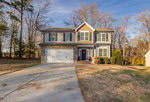 Located at 101 Bluegrass Drive in Oxford, this charming two-story house features gray siding, a stone facade, and a welcoming red front door adorned with a wreath. A concrete driveway leads to a garage on the left, all surrounded by lush trees and grassy areas under a clear, sunny sky.