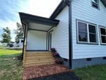 A small white house at 415 Bullock Street boasts a covered entryway with wooden steps and a brick path. Potted flowers adorn the steps, while black trim accentuates the windows and doors. The grassy lawn in this charming Oxford setting is framed by trees in the background.