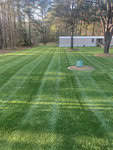 A neatly mowed lawn with visible stripes is surrounded by trees at 2126 Johnson Creek Farms, Oxford. In the distance, a white mobile home is partially visible. A circular mulch area with a green structure stands prominently in the middle of the lawn.