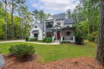 A two-story white house with black trim sits amidst a lush green lawn and surrounding trees. A small American flag is displayed by the front porch, and a paved driveway curves to the side. The sky is partly cloudy.
