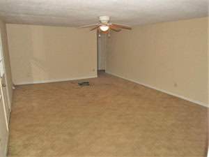 An empty room with beige carpet and walls, featuring a ceiling fan with lights in the center. Near the back wall, a doorway leads to another area. Coiled cables are visible on the floor, and there is a window on the left side.
