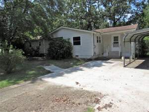 A single-story house with white siding is surrounded by trees. There is a concrete driveway leading to a covered carport on the right. The yard is mostly grass with some bushes near the home. A small set of steps leads to the front door.