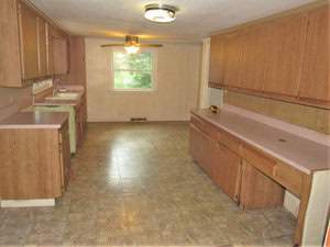 A spacious kitchen with wooden cabinets, light countertops, and a linoleum floor. A window allows natural light in, and a ceiling fan is installed above. The room has a warm, vintage appearance.