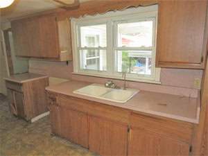 A kitchen with wooden cabinets, a single-basin sink, and a window above the sink. The countertops are a light pink color, and the floor has a patterned design. There's an empty space where a stove might fit.