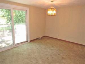 A bright, empty room with beige walls and carpet, featuring a sliding glass door leading to an outdoor area. A golden, hexagonal chandelier hangs from the ceiling, casting a warm glow over the space.