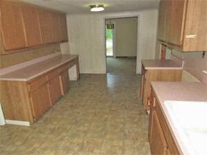 A narrow kitchen with brown wooden cabinets and pink countertops. The floor is covered with patterned linoleum tiles. A doorway leads to another room with a glimpse of a window and light fixture.