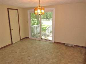 A room with beige carpet and walls features a glass sliding door that opens to a wooden deck with a view of a grassy yard and trees. A gold chandelier hangs from the ceiling, and there's a closed wooden door on the left.