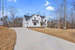 A modern white house with a gray roof sits on a spacious lawn at 997 Browning Place, surrounded by leafless trees. A long paved driveway leads to the entrance, with neighboring houses in the background under Youngsville's clear blue sky.