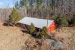 Aerial view of a red barn with a gray metal roof at 579 Walnut Grove Road in Oxford, surrounded by leafless trees and evergreen shrubs. It sits on a grassy field, with a gravel pile nearby and the backdrop of dense and sparse woodland.