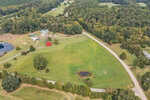 An aerial view of Rougemont reveals a lush green landscape with a winding road leading to 5685 Hobgood Road. A small pond and red barn are nestled among the grassy areas, surrounded by dense woodlands and scattered buildings, crafting a picturesque rural scene.