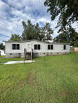 A one-story mobile home with a white exterior and several windows is nestled on a grassy lawn at 109 Pecan Street, Creedmoor. Wooden steps lead to the central front door, set against a backdrop of trees under a partly cloudy sky.