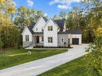 Located at 3912 Sage Court, this expansive two-story white house in Wake Forest boasts stone accents, a black roof, and a spacious driveway. It is surrounded by a manicured lawn and tall trees under a clear blue sky.