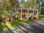 A two-story brick colonial house at 227 Salem Road features tall white columns, surrounded by the lush greenery of Oxford. The driveway gracefully curves in front of the entrance, where sunlight filters through the branches, casting gentle shadows on the lawn and pavement.