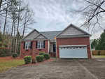 A one-story brick house on 1120 Corrina Road, with a gray roof and a white double garage door. It features black shutters and a small front porch. The concrete driveway is framed by tall trees, under an overcast Wake Forest sky.