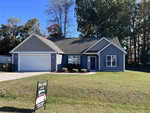 A blue single-story house at 303 Keeneland Drive, Oxford, features a gray roof, two-car garage, and a well-kept lawn. It is surrounded by tall trees and boasts a "New Listing" real estate sign in the front yard.