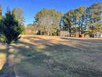 A sunny open field in Franklinton, with patches of grass and scattered trees casting long shadows. A small red shed stands on 3096 Cannady Mill Road, framed by a clear blue sky.