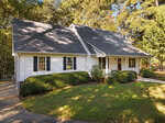 Nestled in a wooded area under a clear sky, this charming white house on 4126 Samuels Court boasts a dark gray gabled roof and black shutters. Surrounded by lush green bushes and trees, it features a welcoming front porch with white railings and a curved driveway typical of Oxford elegance.