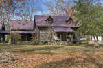 A rustic log cabin with a metal roof, situated at 7561 NC 15 Highway in Oxford, is surrounded by fall foliage. The two-story structure boasts a covered porch and dormer windows. Leaf-covered ground and bare trees suggest late autumn in this rural setting, where a wooden chair sits in the yard.